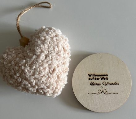 Ein weiches, beiges Herz aus Stoff neben einem runden Holzschild mit Gravur.