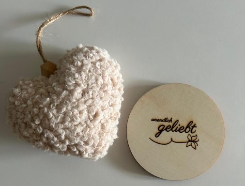Weiches, beiges Herz aus Wolle neben einem runden Holzstück mit der Aufschrift "geliebt".